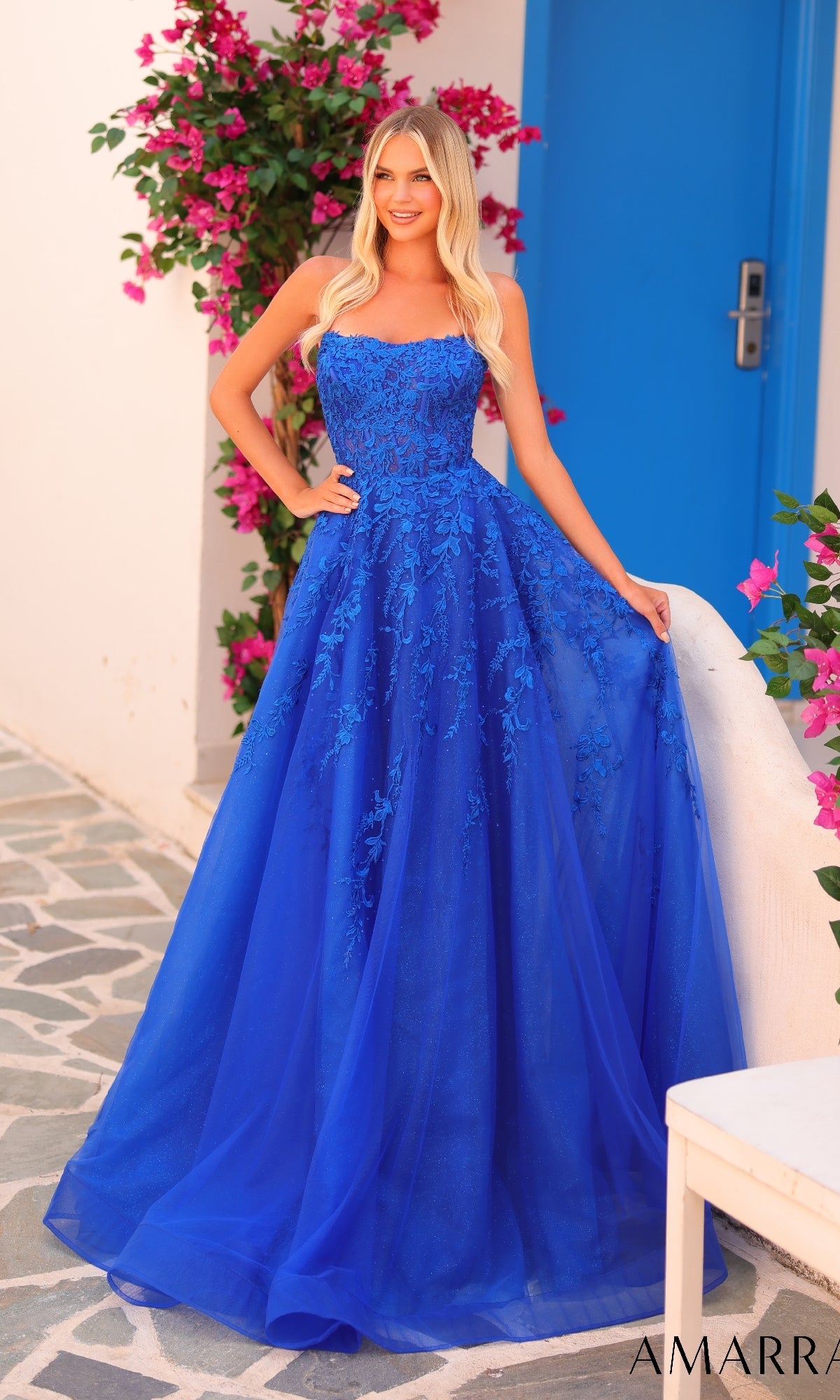 A woman in the Amarra 88258 long prom dress, bright blue with floral lace, stands by a white wall and blue door, surrounded by pink flowers.